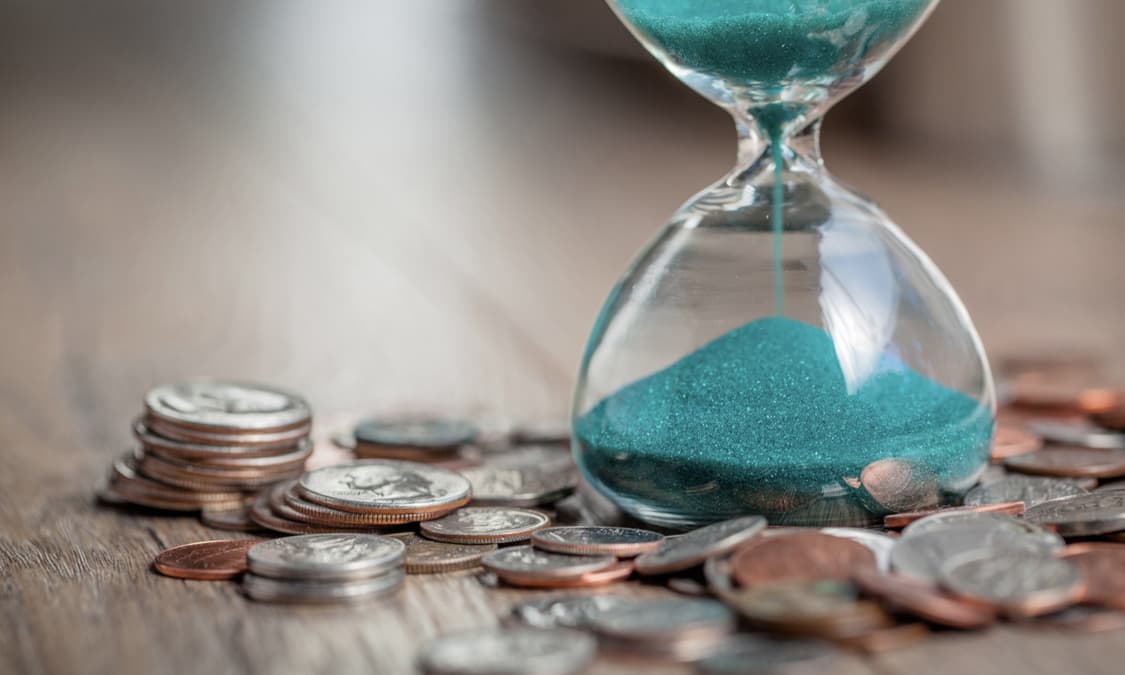 An hour glass filled with teal sand resting on a table full of coins.