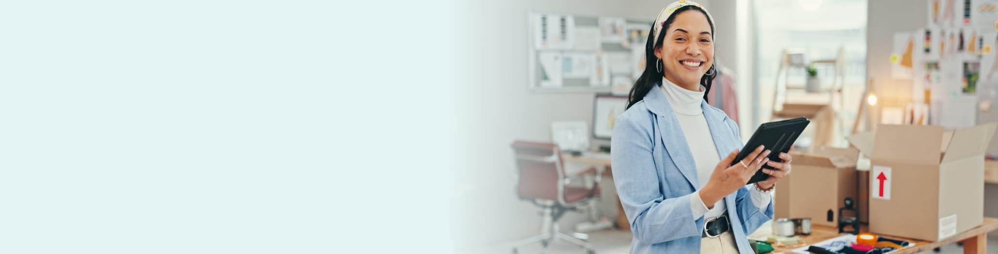 Female small business owner smiling while holding a tablet in a boutique workspace with shipping boxes and merchandise in the background.