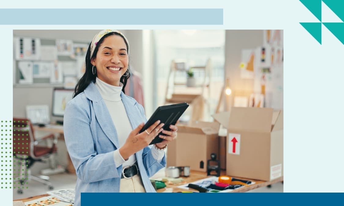 Female small business owner smiling while holding a tablet in a boutique workspace with shipping boxes and merchandise in the background.