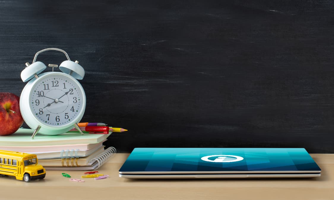 An up close classroom scene with a laptop, books, pencils, and clock on top of a desk with a chalkboard in the background.