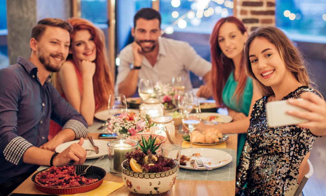 A group of smiling friends sitting at a dinner table taking a selfie.