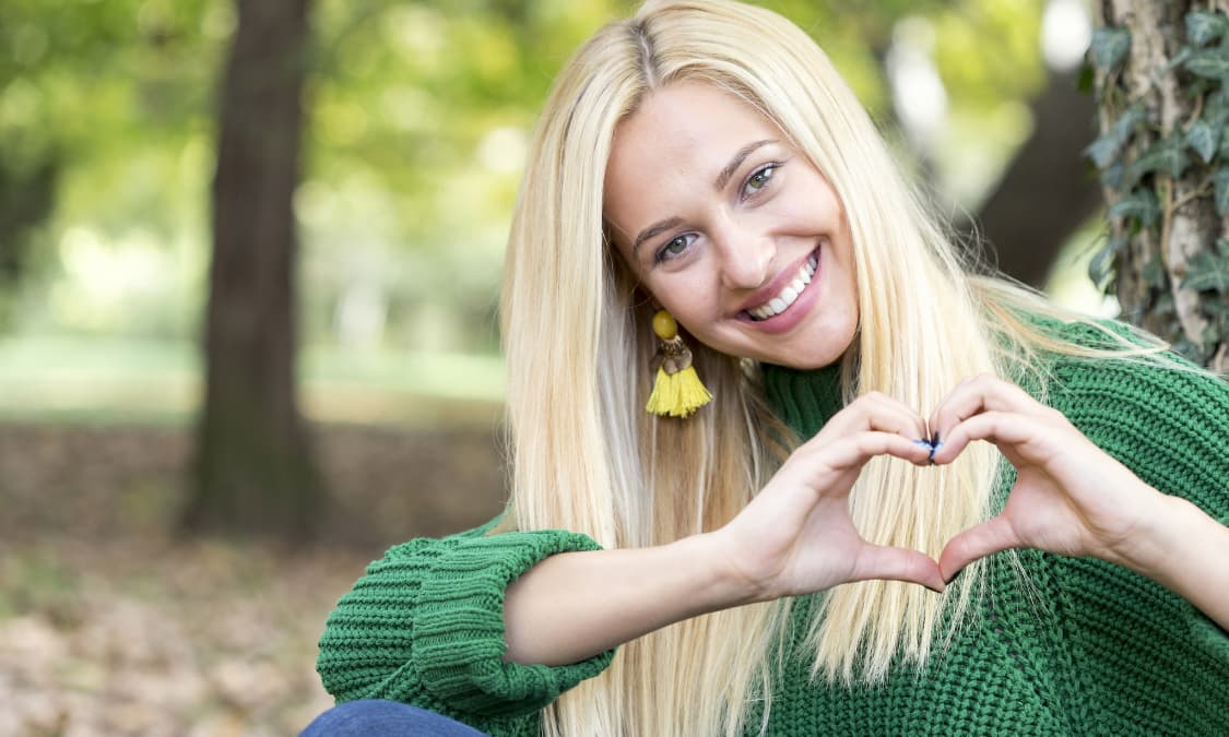 A smiling blonde woman making the shape of a heart with her hands.